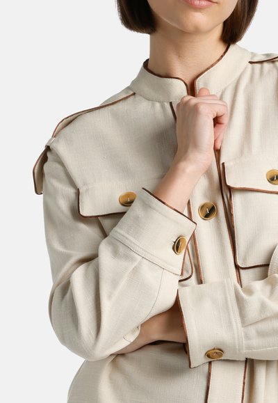 Beige, pitkähihaisessa takissa on korkea kaulus, ruskeat reunat ja kultaiset napit. Siinä on kaksi rintataskua ja rento istuvuus.