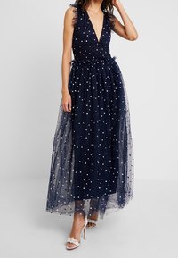 Tumma sininen maksimekko, jossa on V-pääntie, ilman hihmoja ja röyhelökoristeita. Mekossa on tiivis yläosa, läpikuultava pilkkukuosisin päällinen ja lentävä hame.