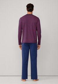 Hombre de pie descalzo, vistiendo una camisa de rayas azul marino y roja de mangas largas, con pantalones lisos azul marino, frente a un fondo liso.