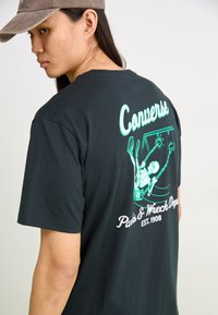 Camiseta de algodón negra con un gráfico verde de un esqueleto en un columpio, con "Converse" y "Parks & Wreck Dept." en texto blanco.