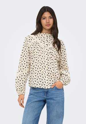 Jeune femme aux longs cheveux foncés portant un chemisier crème à fleurs avec des détails en dentelle et un jean bleu, posant avec une main dans la poche.