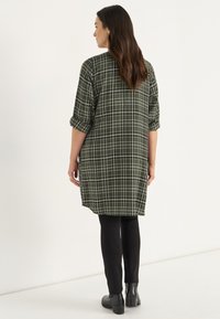 Grüne karierte Tunika mit hochgerollten Ärmeln, die hinten länger ist und seitliche Schlitze hat. Kombiniert mit schwarzen Leggings und Ankle Boots.
