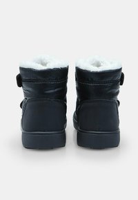 Juodos žiemos botai su balta pūkine pamušalu, matomi iš nugaros, parodantys plokščius guminius padus ir kulkšnies aukščio paminkštintus viršūnius.