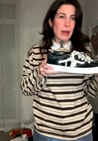 Person mit gestreiftem Hemd hält einen schwarz-weißen Sneaker mit übertrieben weißen Schnürsenkeln, steht drinnen in der Nähe eines Plakats und Schuhkartons.