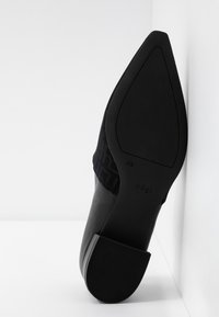 Chaussure à talon noir à bout pointu inclinée verticalement contre un mur d’angle blanc montrant une semelle lisse et un talon bloc.
