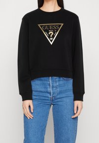 Sudadera negra corta de mezcla de algodón, con un gran logo triangular dorado y texto en el frente; combinada con jeans azules de talle alto.