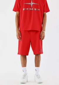 Rode sportoutfit bestaande uit een boxy rode t-shirt met zilveren logo en bijpassende rode shorts. Beide kledingstukken zijn gemaakt van een gladde, lichte stof.