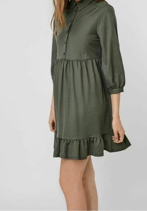 Femme portant une robe ample vert olive avec un col, un devant boutonné, des manches trois-quarts bouffantes et un ourlet à volants.