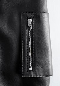 Gros plan d'une manche de veste en cuir noire avec un détail de fermeture éclair métallique argentée verticale cousu dessus.
