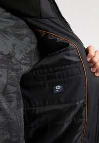 Interior de una chaqueta negra acolchada con un acabado de tela suave, que presenta un forro gris estampado, ribetes naranjas y una etiqueta de marca.