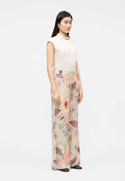 Γυναίκα που στέκεται, φορόντας ένα μπεζ τοπ χωρίς μανίκια και φαρδιά παντελόνια με πολύχρωμο λουλουδάτο και αφηρημένο μοτίβο, καφέ παπούτσια φαίνονται κάτω.
