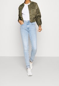Veste bomber vert olive, t-shirt blanc ajusté, jean taille haute bleu clair et baskets blanches, présentant une tenue décontractée.