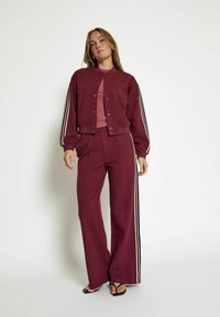 Burgundy tracksuit featuring a bomber-style jacket with snap buttons and striped accents on sleeves, paired with wide-leg pants. Smooth fabric.