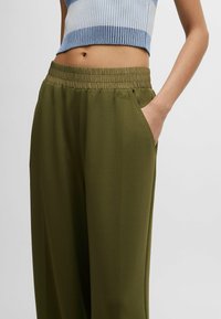 Olivgrüne, elastische Taillen-Hose mit lockerer Passform, ausgestattet mit seitlichen Taschen und dezentem Branding in tonal eingestickter Schrift. Glatte Stofftextur.