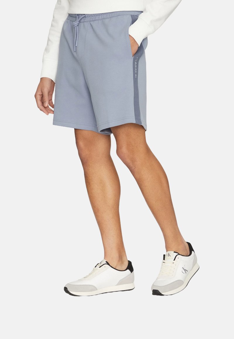 Pantalones cortos azul claro hechos de tela suave, con una cinturilla elástica y un cordón, y bolsillos laterales. Combinados con zapatillas blancas y negras.