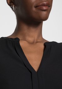 Blouse noire à col en V avec une texture douce et lisse. Elle présente des bords d'épaule arrondis et un éclat subtil, mettant en avant un design minimaliste.