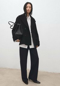 Vrouw met een zwarte double-breasted jas, wit overhemd, wijde broek met krijtstreep, zwarte puntige schoenen en een zwarte handtas tegen een witte muur.