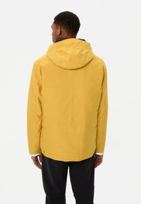 Geel waterdichte jas met capuchon, gladde textuur, verstelbare manchetten en een rechte snit. Achteraanzicht, gecombineerd met zwarte broek.