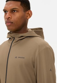 Brauner Kapuzenpullover aus strukturiertem Material, mit einem Reißverschluss, einer Kapuze und einem Logo auf der Brust und dem Ärmel. Schlichtes, sportliches Design.
