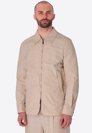 Uomo con la barba che indossa una giacca beige con zip, colletto e tasche laterali, in piedi dritto davanti a uno sfondo semplice.