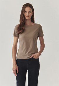 Hnědý top s krátkým rukávem a kulatým výstřihem, vyrobený z hladkého materiálu, v kombinaci s tmavými džíny. Modelka stojí s jednou rukou na boku.