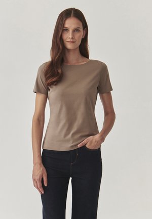 Haut marron à manches courtes avec un col rond, fabriqué dans un tissu lisse, associé à un jean noir. Le mannequin se tient avec une main sur la hanche.