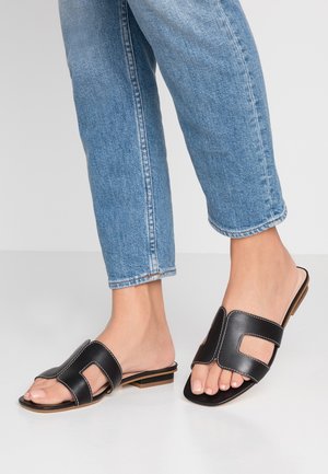 Sandales à glissière en cuir noir avec un design découpé, bout ouvert, semelle plate et couture contrastante, assorties à un jean bleu retroussé.