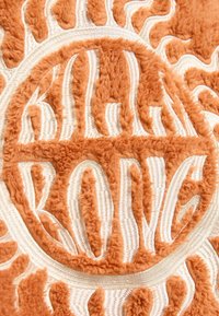 Tissu orange texturé avec une broderie en relief de motifs de soleil, des lignes d'accentuation blanches et des lettres stylisées dans un design circulaire.