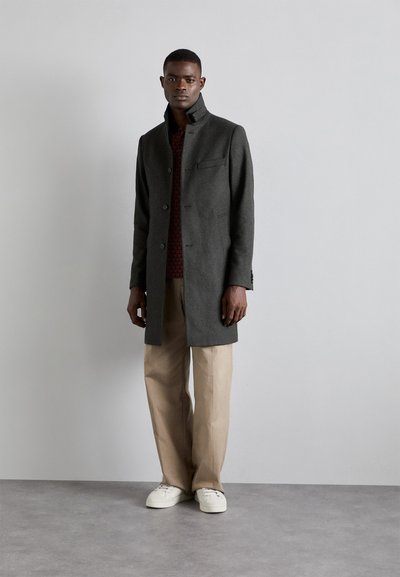 Gray wool coat with a high collar, front buttons, and a chest pocket, worn over a red patterned shirt and cream trousers; casual footwear.