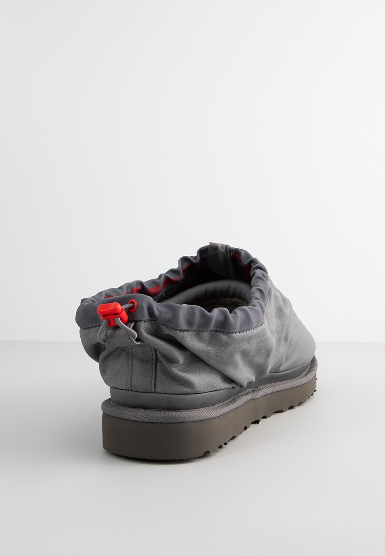 Zapato gris sin cordones con parte superior de tela fruncida, acento rojo, textura suave y cuello acolchado. Suela de goma con un patrón texturizado para mayor agarre.