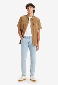 Short-sleeve brown button-up shirt, white t-shirt underneath, light blue jeans, white sneakers with brown soles, standing against a white background.