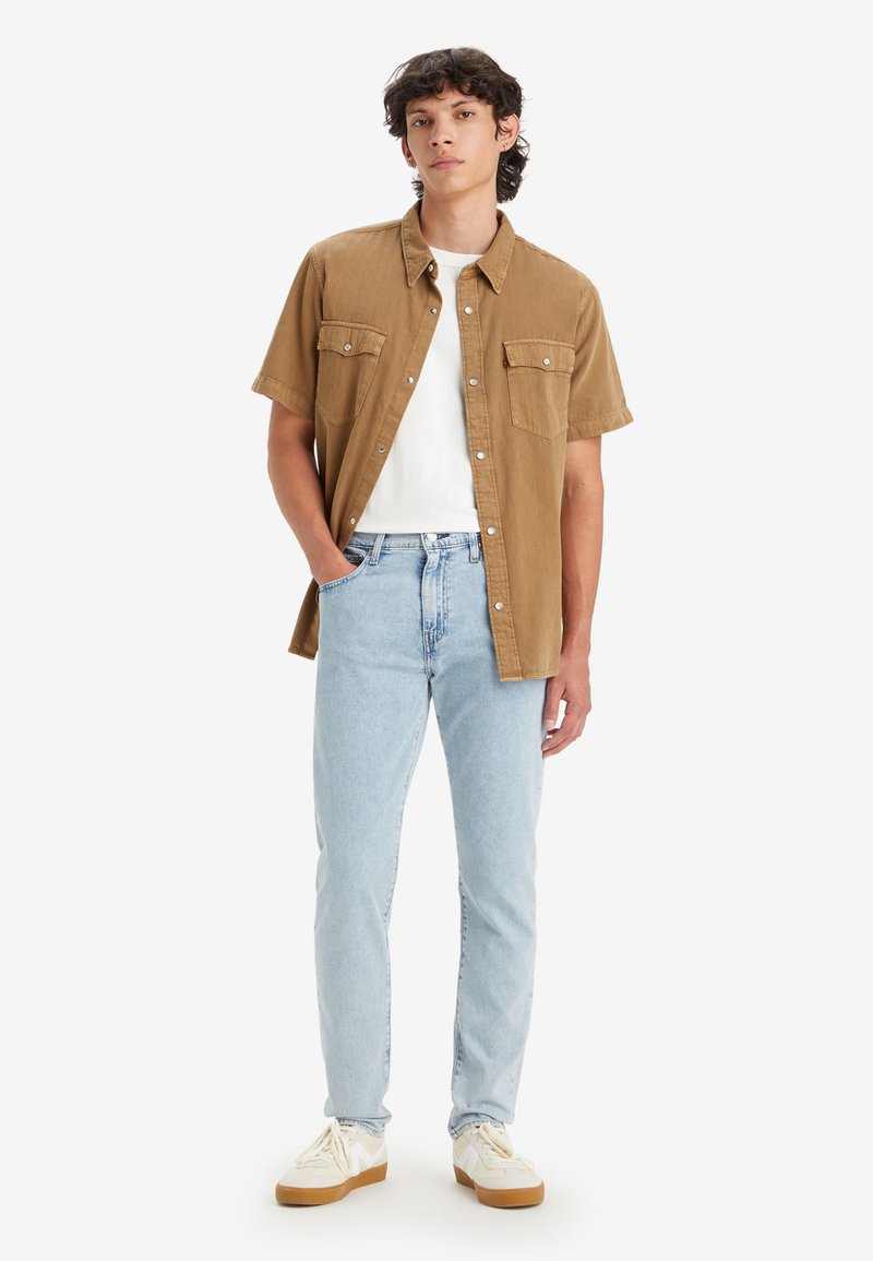 Short-sleeve brown button-up shirt, white t-shirt underneath, light blue jeans, white sneakers with brown soles, standing against a white background.