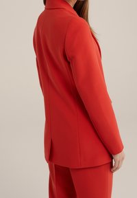 Červený šitý blazer s vystavěným designem, se zářezovým límcem a rozparkem vzadu. Vyroben z hladké, mírně texturované látky.