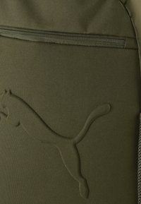 Sac à dos vert olive en tissu texturé, avec un logo Puma en relief bien visible et une poche zippée à l'avant.