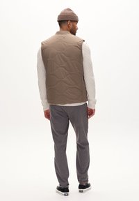 Quilted beige puffer vest with a high collar, worn over a textured white sweater and gray trousers, accompanied by black sneakers and a knit beanie.