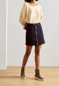 Světle béžový pletený svetr s diamantovým vzorem a širokými rukávy, kombinovaný s námořnickou áčkovou sukní s předním zipem a dvěma kovovými knoflíky. Šedé kotníkové boty na platformě.