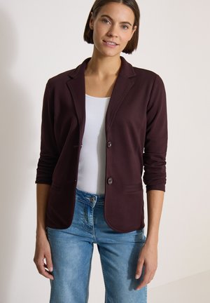 Femme aux cheveux courts marron portant un blazer bordeaux foncé sur un haut blanc et un jean bleu, debout devant un fond clair.