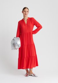 Robe rouge à manches longues à volants avec décolleté en V et corsage plissé. Le mannequin tient un sac argenté et porte des chaussures plates métalliques. Texture lisse.