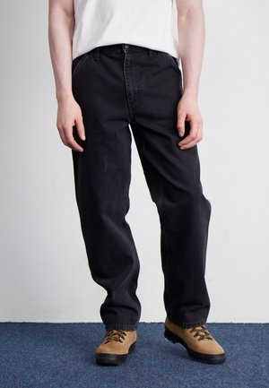 Persoon die losse zwarte jeans, beige wandelschoenen en een wit T-shirt draagt, staand op een blauw tapijt tegen een effen witte muur.