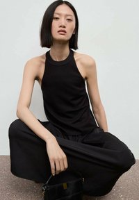 Top halter nero senza maniche abbinato a pantaloni neri fluidi. La modella tiene una piccola borsa nera con un accento dorato, posizionata su uno sfondo neutro.