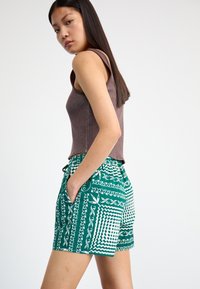 Top de exemplu bej cu dungi, asortat cu pantaloni scurți verzi cu un model geometric alb și buzunare. Materialul are o textură netedă.