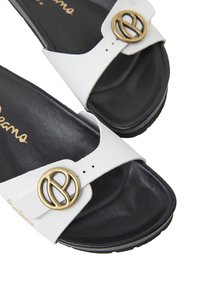 Des sandales blanches à semelle intérieure noire rembourrée et une boucle circulaire dorée proéminente sur la bride. Le design est minimaliste et épuré.