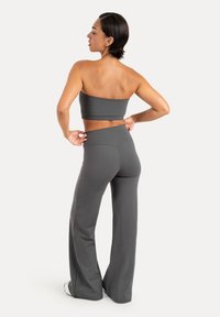 Femme ajustant la taille d’un pantalon large gris foncé, portant un crop top bustier assorti et des baskets blanches, debout sur fond blanc.