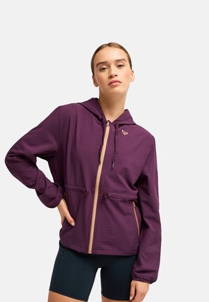 Jeune femme debout portant une veste à capuche violette avec fermeture éclair beige et cordons, associée à un short de cyclisme noir.