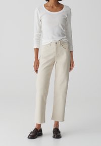 Femme portant un haut blanc à manches longues, un jean beige clair à jambes droites avec ourlets bruts, et des mocassins en cuir marron foncé, debout devant un fond uni.