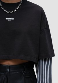 Maglietta nera cropped con logo bianco "WRSTBHVR BERLIN"; presenta maniche corte ed è sovrapposta a una camicia a maniche lunghe a righe.