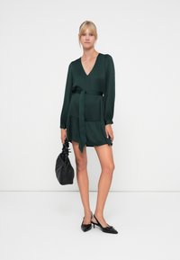 Green dress with a V-neck, long sleeves, and a tie belt. Paired with black slingback shoes and a textured black handbag.