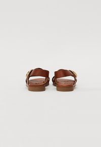 Brown leather sandals featuring a flat sole, dual straps, and a circular gold-tone accent. The design includes open backs and textured insoles.