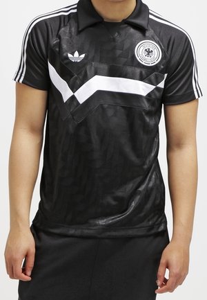 Czarna koszulka piłkarska Adidas z białym paskiem w kształcie szewrona, logo Deutschland Football Band oraz trzema białymi paskami na ramionach, noszona przez osobę.