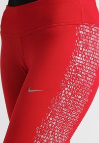 Czerwone legginsy sportowe z gładką strukturą, wysokim pasem i srebrnym logo Nike. Po boku znajduje się pikselowy wzór w kolorze białym.